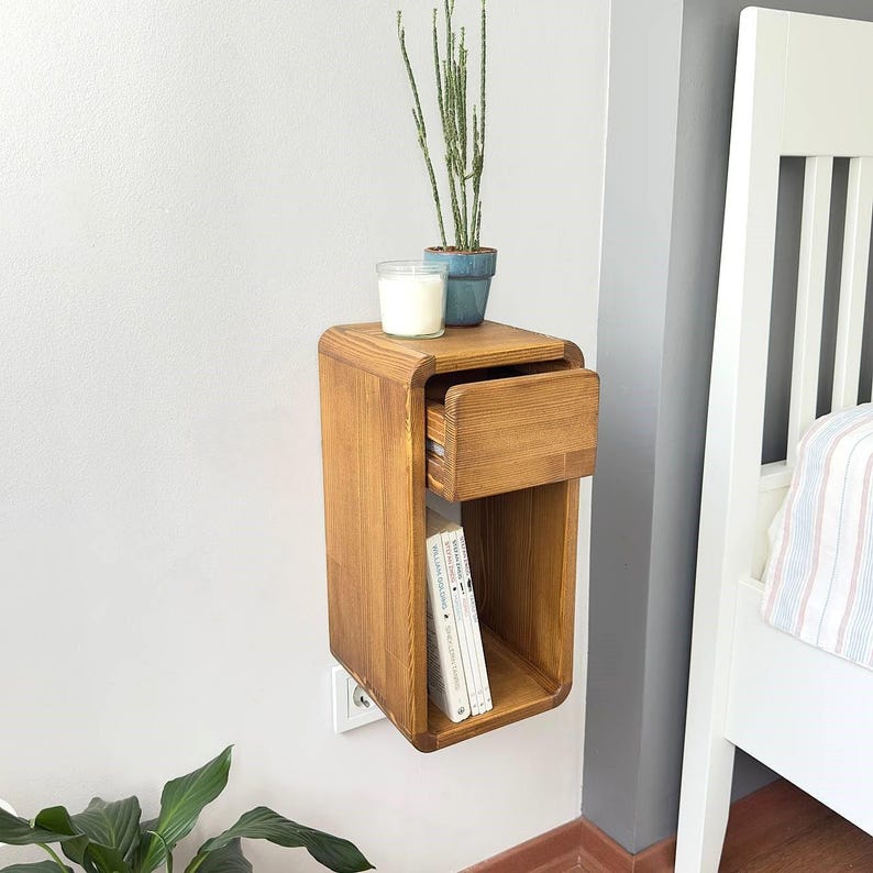 Puede incluir: Una mesita de noche flotante de madera con un caj&oacute;n y una estanter&iacute;a abierta. La mesita de noche es de madera marr&oacute;n claro y est&aacute; montada en una pared gris. Una peque&ntilde;a planta en maceta, una vela y libros est&aacute;n encima.