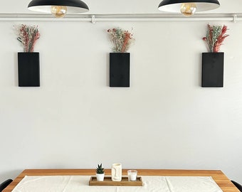 May include: A wooden dining table with a white tablecloth and a small succulent plant in a white pot. There are three black rectangular wall decor pieces with dried flowers in front of them. A white ceramic mug and a small candle are on the table.