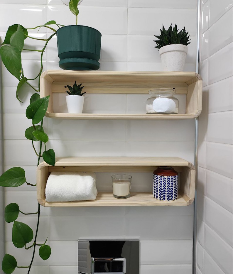 May include: Two wooden shelves with rounded edges, mounted on a white tiled wall. The top shelf has a green potted plant, a small potted succulent, and a glass jar with cotton rounds. The bottom shelf has a white towel, a small glass candle, and a blue and white patterned jar.