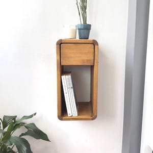 Puede incluir: Estante flotante de madera marr&oacute;n claro con un caj&oacute;n y almacenamiento abierto. Una pila de libros est&aacute; en el espacio abierto. Una peque&ntilde;a maceta azul con una planta y una vela se encuentran encima.