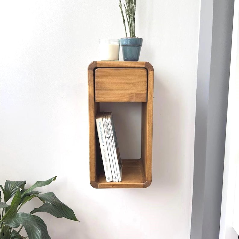 Puede incluir: Estante flotante de madera marr&oacute;n claro con un caj&oacute;n y almacenamiento abierto. Una pila de libros est&aacute; en el espacio abierto. Una peque&ntilde;a maceta azul con una planta y una vela se encuentran encima.