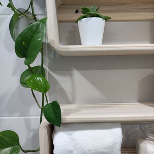 May include: Two wooden shelves with rounded edges, mounted on a white tiled wall. The top shelf has a small green succulent plant in a white pot. The bottom shelf has a white towel and a clear glass jar with white cotton rounds.