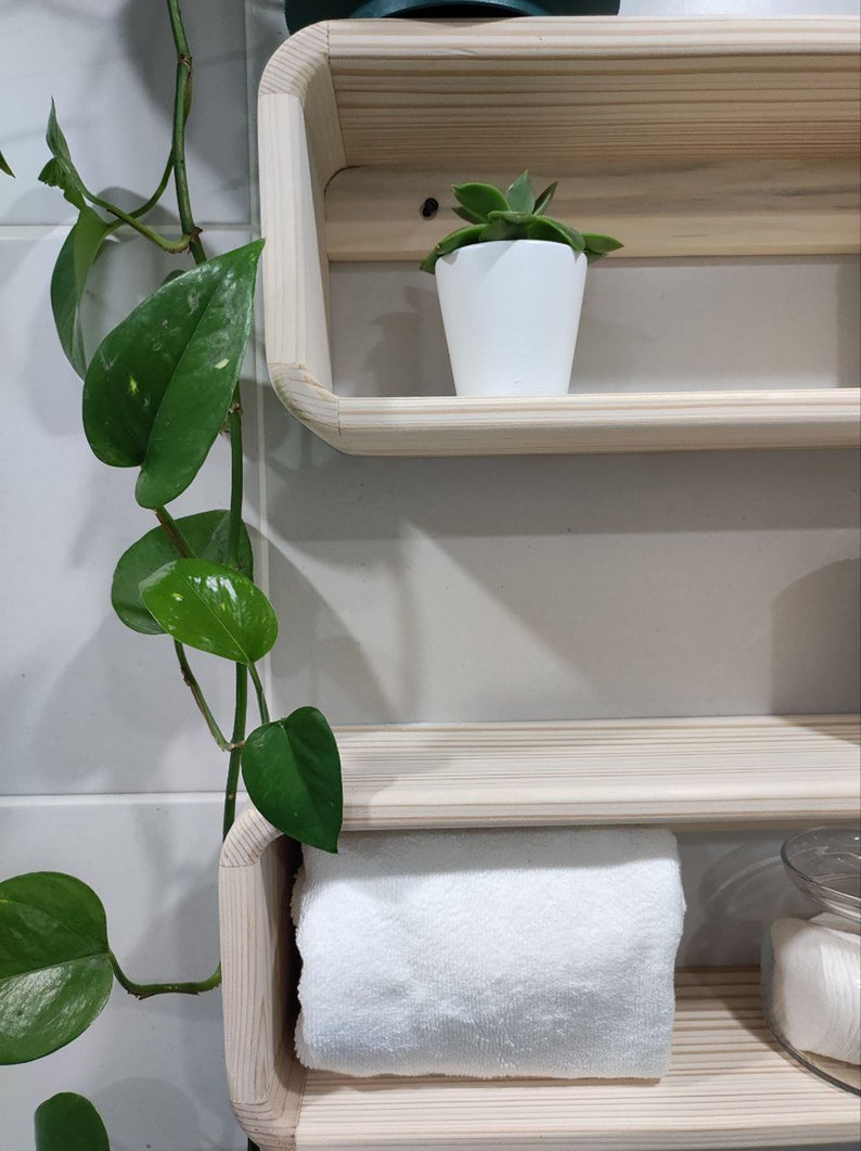 May include: Two wooden shelves with rounded edges, mounted on a white tiled wall. The top shelf has a small green succulent plant in a white pot. The bottom shelf has a white towel and a clear glass jar with white cotton rounds.
