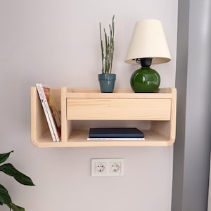 Peut inclure: Une étagère murale en bois clair avec un tiroir, deux étagères et un petit cactus en pot. L'étagère a un bord arrondi et est montée sur un mur blanc. Une lampe verte avec un abat-jour blanc est posée sur l'étagère supérieure. L'étagère a également une pile de livres et un livre noir sur l'étagère inférieure.