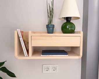 May include: A light wood wall shelf with a drawer, two shelves, and a small potted cactus. The shelf has a rounded edge and is mounted on a white wall. A green lamp with a white shade sits on the top shelf. The shelf also has a stack of books and a black book on the lower shelf.