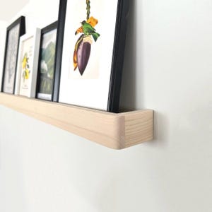 May include: A light-colored wooden floating shelf mounted on a white wall, holding framed art prints. The shelf has a rounded edge. The art includes botanical and landscape prints in black frames.