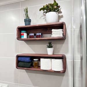 Könnte beinhalten: Zwei Holzregale mit abgerundeten Kanten, die an einer weißen gekachelten Wand montiert sind. Das obere Regal hat eine kleine Topfpflanze, ein Glas und drei kleine Behälter. Das untere Regal hat zwei Toilettenpapierrollen, eine kleine Flasche und zwei gefaltete Handtücher.