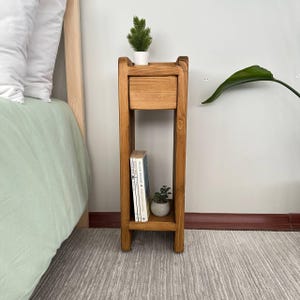 Puede incluir: Una mesita de noche de madera marrón claro con un cajón y un estante. La mesita tiene una pequeña planta en maceta encima y una pequeña planta en maceta y dos libros en el estante. La mesita está al lado de una cama. Altura aproximada 76 cm.