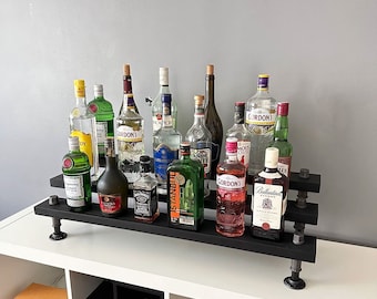 Puede incluir: Una estantería de metal negro con una variedad de botellas de licor, incluyendo ginebra, vodka, ron y whisky. Las botellas están dispuestas en dos filas en la estantería, con algunas botellas delante de otras. La estantería está montada en una pared blanca.