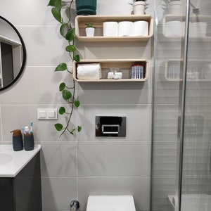 May include: A white bathroom with a toilet, a shower, and two wooden shelves. The shelves are decorated with plants, towels, and other bathroom items.