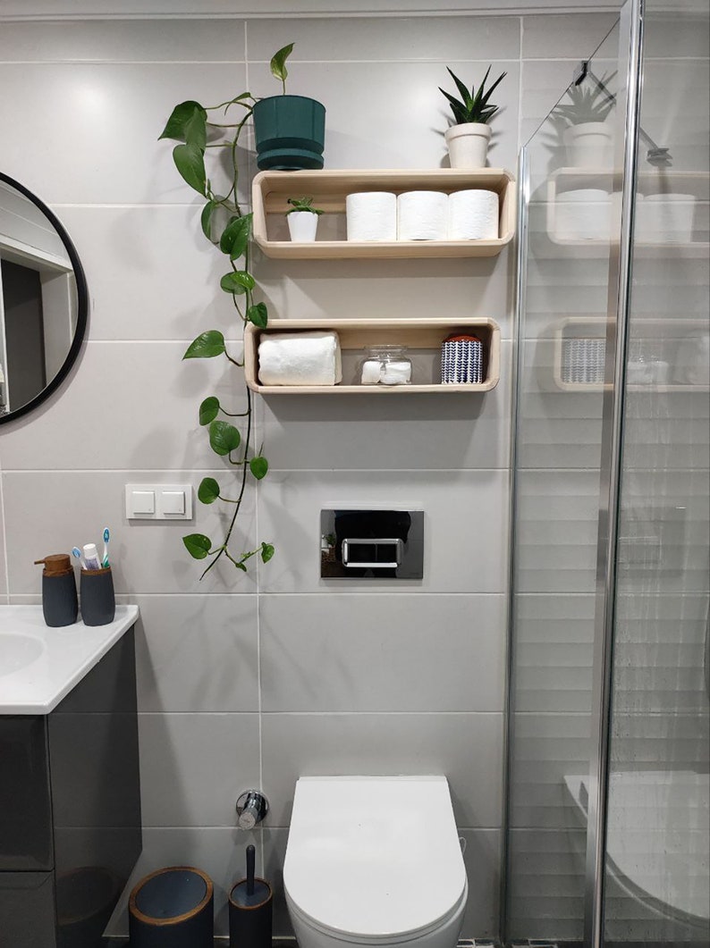 May include: A white bathroom with a toilet, a shower, and two wooden shelves. The shelves are decorated with plants, towels, and other bathroom items.