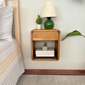 Può includere: Un comodino in legno sospeso con un cassetto e un ripiano aperto. Una lampada verde con paralume color crema è appoggiata sopra, accanto a una piccola pianta in vaso. Il ripiano contiene libri e una piccola succulenta in un vaso di cemento.