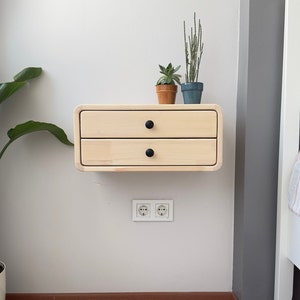 Könnte beinhalten: Ein schwebendes Regal aus hellem Holz mit zwei Schubladen und schwarzen Knöpfen. Das Regal ist an einer weißen Wand montiert und hat zwei Steckdosen darunter. Zwei kleine Topfpflanzen stehen auf dem Regal.