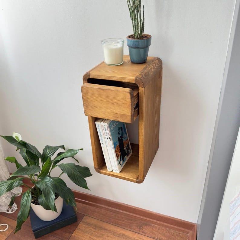 Puede incluir: Una mesita de noche de color marr&oacute;n claro, montada en la pared, con un caj&oacute;n y una estanter&iacute;a abierta. El caj&oacute;n est&aacute; parcialmente abierto, revelando el interior. La estanter&iacute;a contiene dos libros. Una vela en un tarro de cristal y una peque&ntilde;a planta en maceta est&aacute;n encima.