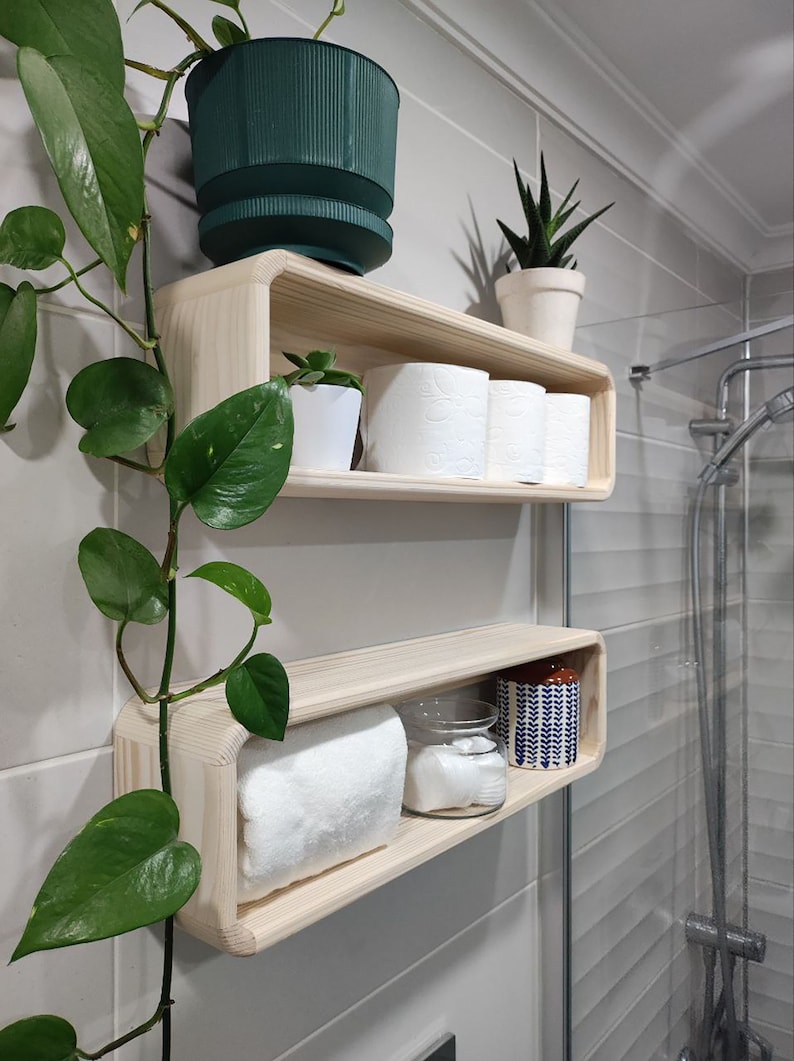 May include: Two wooden shelves with rounded edges, mounted on a white tiled wall. The top shelf holds a green potted plant, three rolls of toilet paper, and a small white potted plant. The bottom shelf holds a white towel, a glass jar, and a blue and white patterned jar.