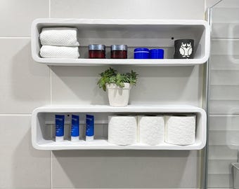 May include: Two white, rectangular shelves mounted on a tiled wall. The top shelf holds folded white towels, jars, and a black owl-themed container. The bottom shelf displays blue tubes, a potted plant, and rolls of toilet paper.