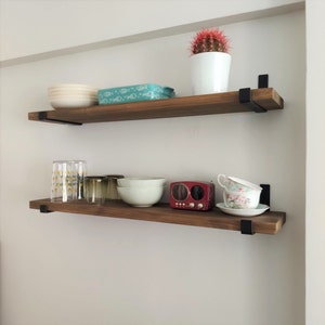 Puede incluir: Dos estantes de madera con soportes de metal negro, que sostienen varios artículos, incluyendo platos, vasos, una radio pequeña y un cactus en maceta.