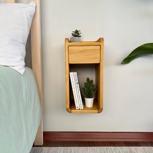 May include: A wooden wall-mounted bedside table with a drawer and a shelf. The bedside table is light brown and has a curved front edge. There are books and a small potted plant on the shelf.