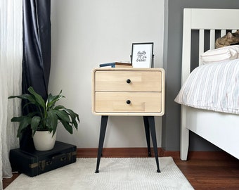 May include: A light wood nightstand with two drawers and black knobs. The nightstand has black metal legs and rounded corners. A framed print with the text "Beauty is in the beholder" sits on top. A plant and a suitcase are in the foreground.