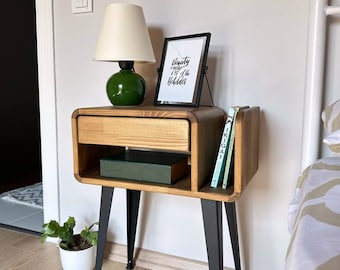 Puede incluir: Una mesita de noche de madera con un cajón, un estante y un soporte para libros. La mesa tiene patas de metal negro y una lámpara verde con una pantalla blanca. Una impresión enmarcada con el texto "La belleza viene de adentro" está sobre la mesa.