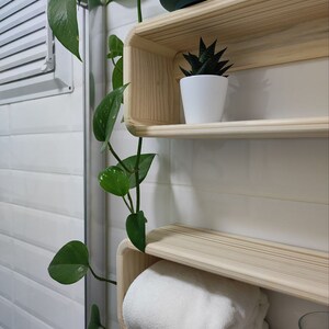 May include: Two wooden shelves with rounded edges, mounted on a white tiled wall. The top shelf has a small potted succulent plant, and the bottom shelf has a white towel folded on it.