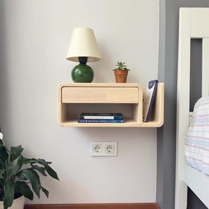 Könnte beinhalten: Ein schwebendes Nachttischchen aus hellem Holz mit einer Schublade und einem Regal. Eine grüne Lampe mit weißem Schirm steht auf dem oberen Regal. Eine kleine Topfpflanze steht auf der Schublade. Der Nachttisch ist an einer weißen Wand neben einem weißen Bett befestigt.