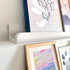 Op de afbeelding: Wit houten plank met een gebogen rand, met ingelijste kunstwerken. De kunstwerken omvatten een zwart-witte bloemenprint, een kleurrijk abstract schilderij en een zwart-witte print met een geometrisch ontwerp.