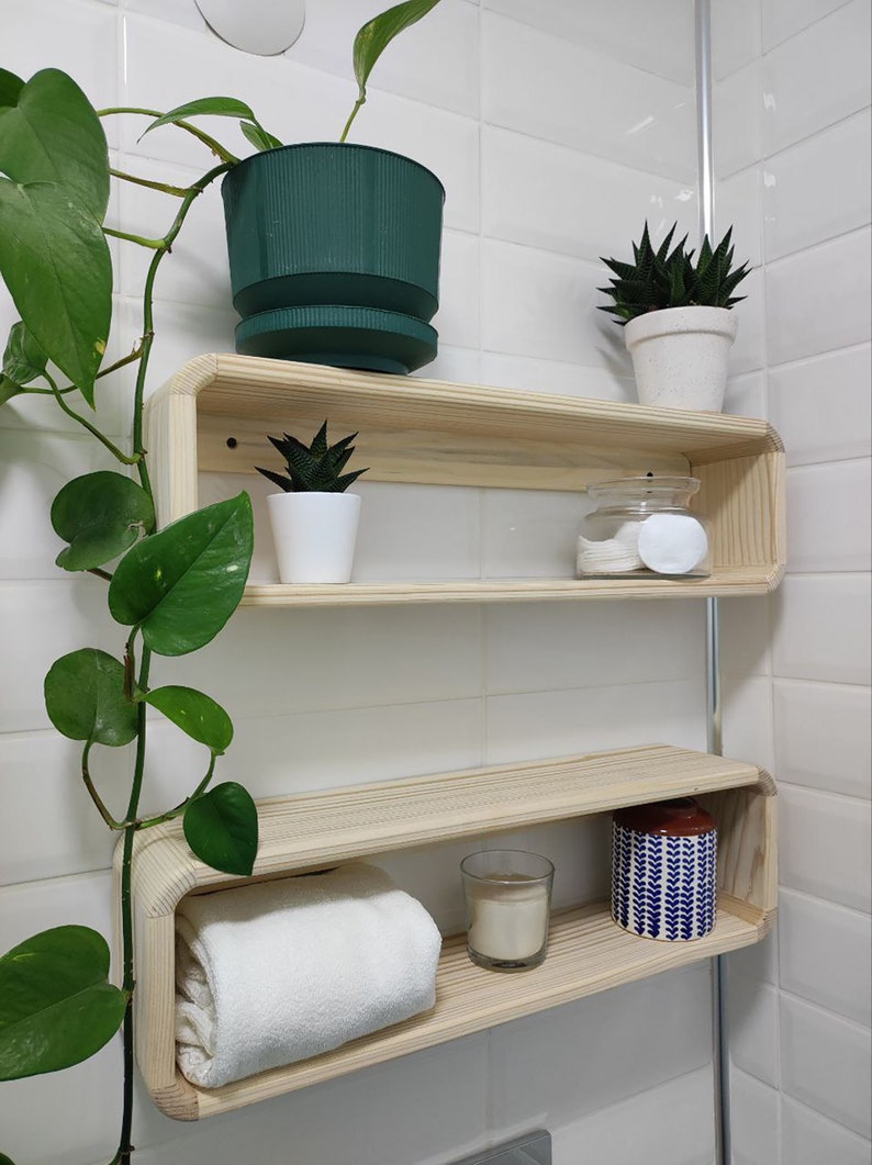 May include: Two wooden shelves with rounded edges, mounted on a white tiled wall. The top shelf has a green potted plant, a white potted succulent, and a glass jar with cotton rounds. The bottom shelf has a white towel, a white candle in a glass jar, and a blue and white patterned jar.