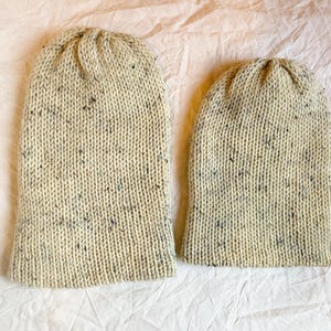 Könnte beinhalten: Zwei cremefarbene Strickmützen mit Flecken in Dunkelgrau und Blau. Die Mützen sind aus einem strukturierten Garn gefertigt und haben einen gerippten Bund. Die Mützen werden auf einer weißen Oberfläche gezeigt.