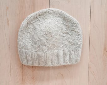 Pode incluir: Um gorro de malha cinza claro. O gorro tem uma aba canelada e uma coroa texturizada com padrão. O gorro é feito de um material macio e quente, ideal para o frio. O gorro é mostrado em uma superfície de madeira clara.