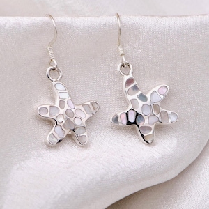 May include: A pair of silver-toned dangle earrings shaped like starfish. Each starfish features a mosaic inlay of iridescent, multi-colored shell fragments set within a polished silver-colored metal frame, suspended from simple silver-toned ear wires.