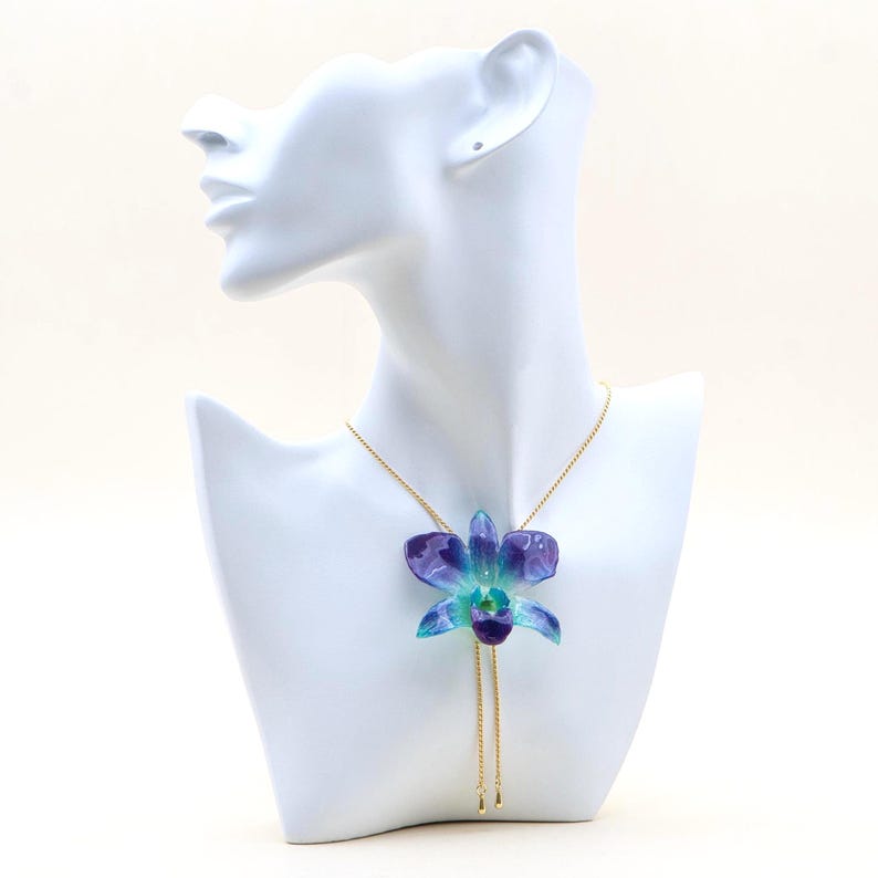 May include: A necklace featuring a pendant shaped like a blue and purple orchid flower. The flower is suspended from a delicate gold chain. The necklace is displayed on a white mannequin bust.