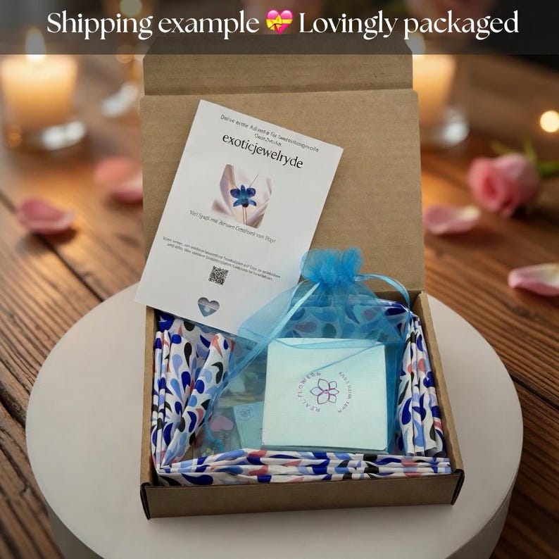May include: An open cardboard box reveals a gift set. Inside, a blue drawstring bag, a small white card with a flower logo, and a folded paper with text are visible. The box is lined with a patterned fabric.