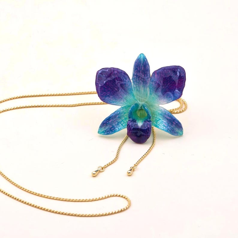 May include: A pendant necklace featuring a detailed, translucent orchid flower in shades of blue and purple. The flower is suspended from a delicate gold-colored chain. The necklace is set against a white background, highlighting the vibrant colors of the floral design.
