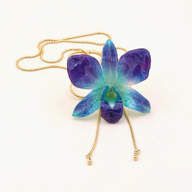 May include: A pendant necklace featuring a blue and purple orchid flower. The flower has a gold chain and two dangling gold chains. The orchid has a gradient of colors from blue to purple.