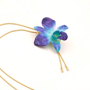 May include: A gold-colored chain necklace with a pendant shaped like a blue and purple orchid flower. The orchid has a gradient of colors, with a light blue center and darker purple edges. The necklace has a delicate, feminine design.