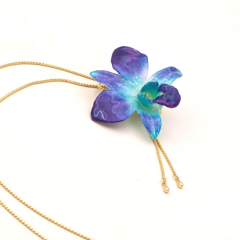 May include: A gold-colored chain necklace with a pendant shaped like a blue and purple orchid flower. The orchid has a gradient of colors, with a light blue center and darker purple edges. The necklace has a delicate, feminine design.