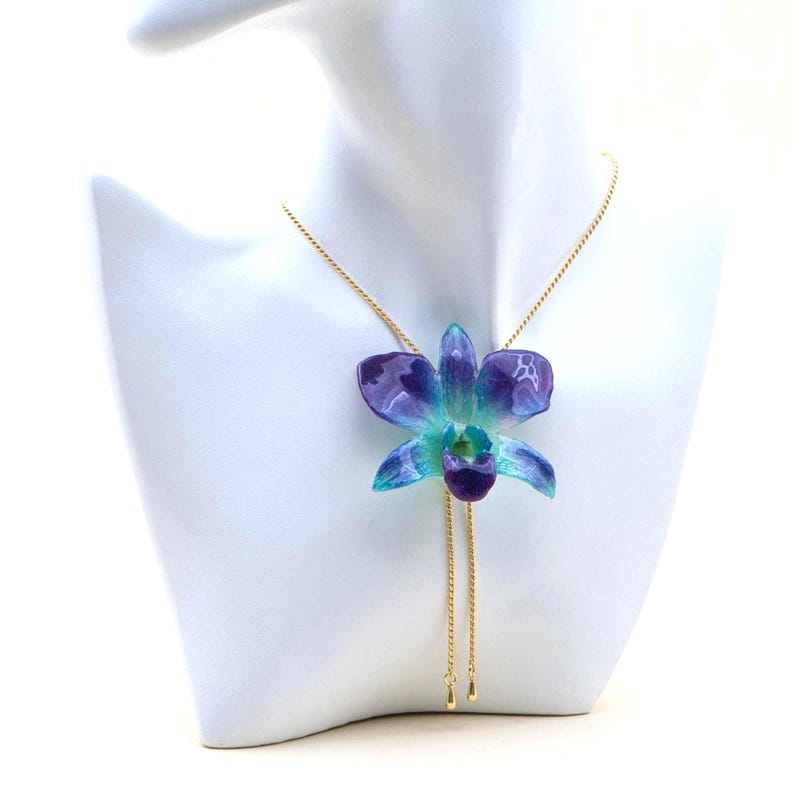Könnte beinhalten: Eine goldfarbene Halskette mit einem Anhänger in Form einer blau-violetten Orchideenblüte. Die Orchidee wird auf einer weißen Mannequin-Büste präsentiert. Die Halskette hat eine zarte Kette und einen Schiebeverschluss.