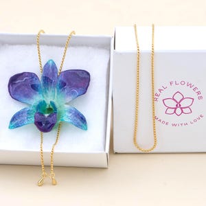 May include: A handcrafted necklace featuring a vibrant blue and purple orchid pendant. The flower is set on a delicate gold chain, presented in a white box. The box has the text "REAL FLOWERS MADE WITH LOVE" and a flower logo.