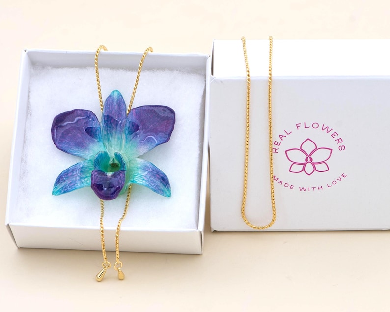 May include: A handcrafted necklace featuring a vibrant blue and purple orchid pendant. The flower is set on a delicate gold chain, presented in a white box. The box has the text "REAL FLOWERS MADE WITH LOVE" and a flower logo.