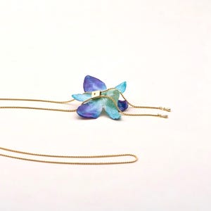 May include: A handcrafted necklace featuring a blue and purple flower pendant with gold-colored chain. The flower has a square gold-colored accent in the center. The necklace is set against a white background.