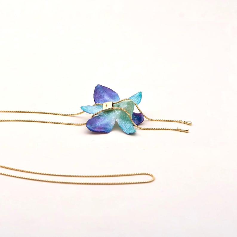 May include: A handcrafted necklace featuring a blue and purple flower pendant with gold-colored chain. The flower has a square gold-colored accent in the center. The necklace is set against a white background.