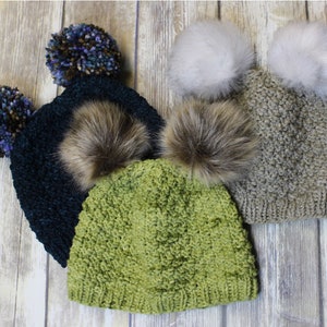 Op de afbeelding: Drie gebreide mutsen met verschillende gekleurde pompoenen. De mutsen zijn groen, blauw en grijs. De pompoenen zijn gemaakt van bont en garen.