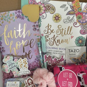 Könnte beinhalten: Eine Geschenkbox gefüllt mit Artikeln zum Thema Glaube und Hoffnung. Die Box enthält ein Malbuch mit dem Titel "Be Still & Know", ein Tagebuch mit den Worten "faith hope", ein flauschiges rosa Haargummi, ein Paar rosa Socken mit den Worten "Let Go + Let God" und andere Kleinigkeiten.
