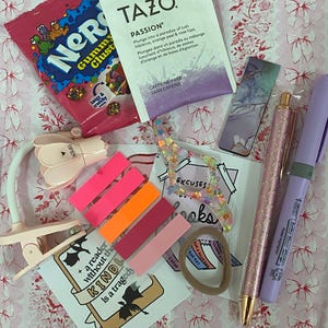 May include: A collection of stationery and treats on a pink floral background. Includes a packet of Nerds Gummy Clusters, Tazo Passion tea, sticky notes, pens, a bookmark, a hair tie, and a reading light.