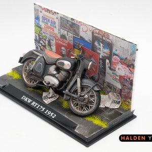 Peut inclure: Modèle de moto DKW RT175 noir et argent de 1952, garée contre un mur recouvert d'affiches vintage. Le modèle est présenté sur une base noire avec de l'herbe verte et une étiquette indiquant "DKW RT175 1952".