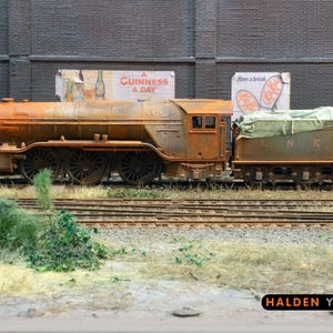 Può includere: Un motore di treno modello arrugginito, consumato e invecchiato con una tela cerata verde che copre la parte posteriore del motore. Il treno è seduto su un binario di treno modello con un muro di mattoni e una pubblicità vintage sullo sfondo. La pubblicità recita "A Guinness a Day".