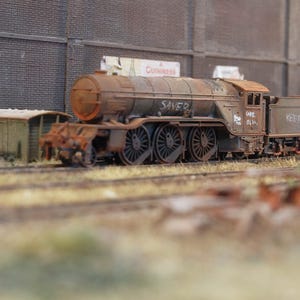 Può includere: Modello dettagliato di una locomotiva a vapore arrugginita sui binari. Le parole "SAVED" e "KEEP" sono visibili sul treno. Il modello è marrone e ha una caldaia cilindrica e più ruote. Un piccolo edificio e un muro di mattoni sono sullo sfondo.