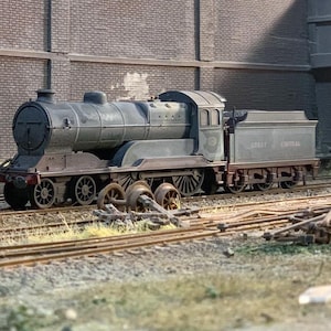 Peut inclure: Maquette détaillée d'une locomotive à vapeur vintage, peinte dans des tons de gris et de vert, avec l'inscription "GREAT CENTRAL" visible sur le côté. Le modèle est posé sur une voie ferrée miniature, avec des morceaux de voie et des débris supplémentaires à proximité.
