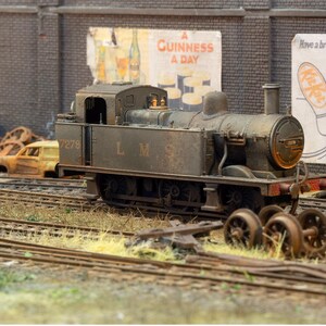May include: A rusty green model train engine with the number 7279 and the letters LMS on the side. The engine is sitting on a set of train tracks with brown grass and dirt around it. There are two posters in the background, one for Guinness and one for Kit Kat.